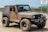 A muddy off-road vehicle, likely a Jeep, is parked on a dirt or sandy path. The vehicle is covered in dirt, indicating it has been used in rough terrain. It has large, rugged tires suitable for off-roading and additional equipment on the front, such as a winch and extra lights. The surrounding area is sparse with some greenery and a sign in the background.
