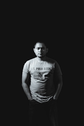 A person stands with hands in pockets against a dark background, wearing a t-shirt with text and a graphic. The lighting casts shadows, creating a dramatic atmosphere.
