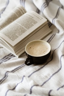 An open book with printed text lies on a soft, textured fabric with blue stripes. Next to the book, a black cup filled with a frothy beverage rests, creating a cozy and relaxed atmosphere.
