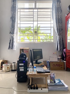 A home office setup on the floor in a well-lit room with a large window framed by blue curtains. Outside, palm trees are visible. Inside, there is a computer setup with a monitor, keyboard, and CPU, along with a bottle of hand sanitizer and a mask. A red cabinet is on the right, and there are boxes and household items like a kettle and rice cooker nearby.