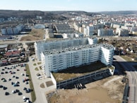 Aerial view of a residential area featuring large, modern apartment buildings with white facades and multiple balconies. The structures are surrounded by parking lots with numerous parked cars. The background includes a hilly landscape with scattered rows of houses and urban infrastructure.