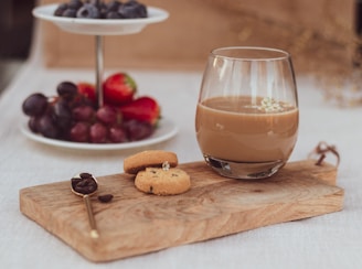 clear glass cup with brown liquid beside red berries on brown wooden chopping board