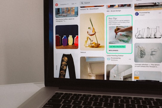 A laptop is displaying a Pinterest-like interface with various images and advertisements. The screen includes colorful vases, a watercolor painting tutorial, abstract art, and a kitchen interior. A small portion of the laptop's keyboard is visible at the bottom.