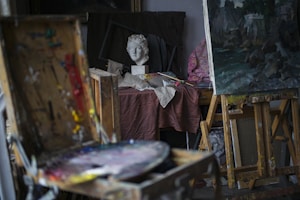 An artist's studio filled with various painting equipment including a palette with paint, brushes, and easels. A painted canvas is placed on one easel, and a sculpted head bust is situated on a table covered with a cloth. The setting is cluttered, indicative of an active, creative space.