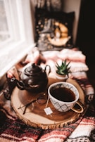 A cozy tea setup with a teapot, cups, and natural leaves on a wooden tray by a window.