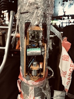 A traffic pole or similar structure with an exposed electronic mechanism is wrapped in plastic and secured with red and white tape. The circuitry is visible, revealing components like a circuit board, a motor, and various wires. The surrounding area is urban, with blurred lights and buildings in the background, creating a nighttime city scene.
