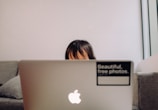 A child partially visible behind a laptop, sitting on a couch. The laptop has an Apple logo and a sticker reading 'Beautiful, free photos.' on its lid. The background is a plain, light-colored wall, and the setting appears cozy and casual.