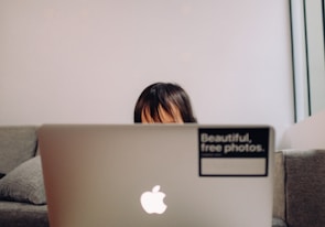 A child partially visible behind a laptop, sitting on a couch. The laptop has an Apple logo and a sticker reading 'Beautiful, free photos.' on its lid. The background is a plain, light-colored wall, and the setting appears cozy and casual.