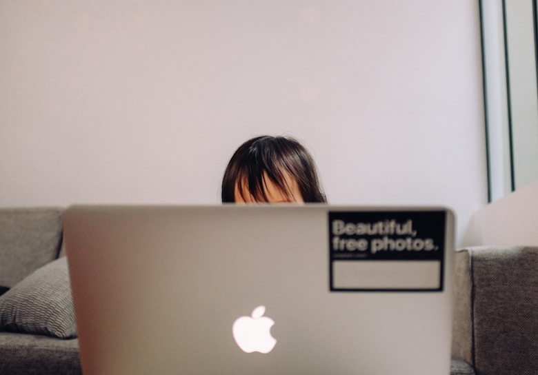 A child partially visible behind a laptop, sitting on a couch. The laptop has an Apple logo and a sticker reading 'Beautiful, free photos.' on its lid. The background is a plain, light-colored wall, and the setting appears cozy and casual.