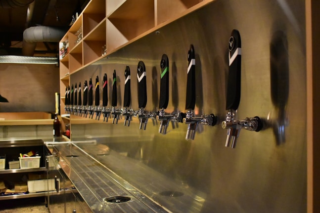 A long row of beer tap handles is mounted on a stainless steel wall in a bar setting. The taps are uniformly aligned, each with a distinct color marking or logo. The bar counter is clean, and shelves above hold various items. The lighting gives a warm, ambient glow to the scene.