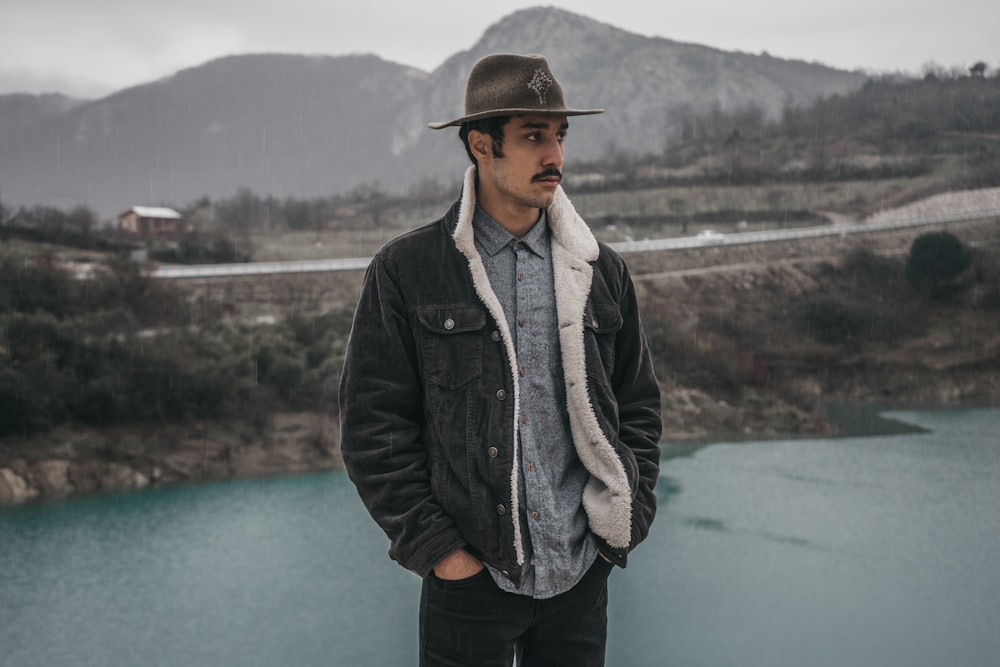 man in black leather jacket and blue denim jeans standing near body of water during daytime