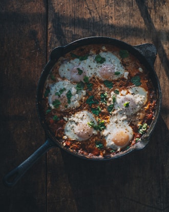 A cast iron skillet filled with a dish composed of eggs poached in a rich tomato sauce, garnished with fresh herbs, resting on a rustic, dark wooden surface. The eggs have runny yolks and are scattered with bits of seasoning and herbs, creating a contrasting texture and color palette. Soft shadows and light hint at a cozy, warm setting.