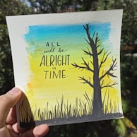 A hand is holding a piece of paper featuring a watercolor illustration with a tree silhouette on the right and blades of grass at the bottom. The background features a gradient of blue, green, and yellow colors. The paper contains motivational text in bold letters stating: 'All will be alright in time.'