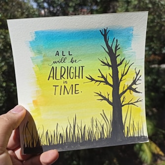 A hand is holding a piece of paper featuring a watercolor illustration with a tree silhouette on the right and blades of grass at the bottom. The background features a gradient of blue, green, and yellow colors. The paper contains motivational text in bold letters stating: 'All will be alright in time.'