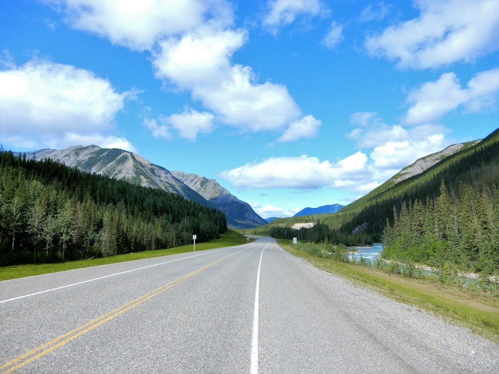 gray concrete road between green trees and mountains under blue sky and white clouds during daytime