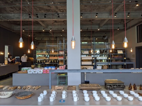 A modern interior space with exposed concrete ceiling, featuring industrial-style pendant lights hanging above a large counter. The foreground displays neatly arranged white cups and various artisanal goods, such as bread baskets and bottles. The background has shelves stocked with products and a few people, one of whom appears to be interacting with a customer.