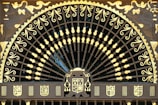 An ornate, semi-circular wrought iron gate features intricate patterns and designs highlighted in gold. The upper section is embellished with a series of curved lines and floral motifs, radiating from a central point, resembling a fan or peacock tail. Surrounding the fan-shaped design, there are artistic depictions of figures and heraldic shields in gold. The overall appearance is regal and lavish, with a strong emphasis on craftsmanship.