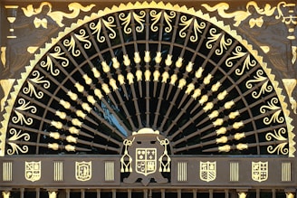 An ornate, semi-circular wrought iron gate features intricate patterns and designs highlighted in gold. The upper section is embellished with a series of curved lines and floral motifs, radiating from a central point, resembling a fan or peacock tail. Surrounding the fan-shaped design, there are artistic depictions of figures and heraldic shields in gold. The overall appearance is regal and lavish, with a strong emphasis on craftsmanship.