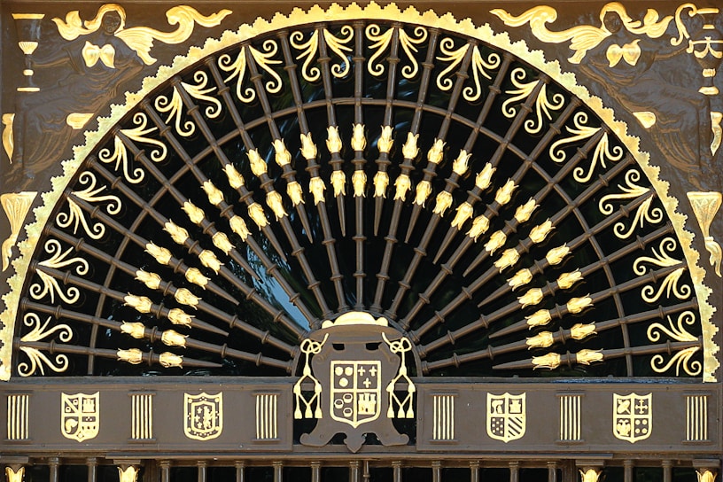 An ornate, semi-circular wrought iron gate features intricate patterns and designs highlighted in gold. The upper section is embellished with a series of curved lines and floral motifs, radiating from a central point, resembling a fan or peacock tail. Surrounding the fan-shaped design, there are artistic depictions of figures and heraldic shields in gold. The overall appearance is regal and lavish, with a strong emphasis on craftsmanship.