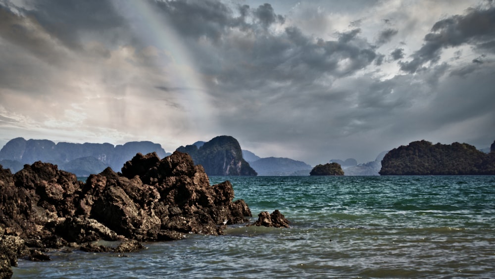 rocky mountain beside body of water under cloudy sky during daytime