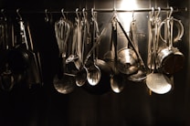 A collection of various kitchen utensils hanging from hooks on a metal rack. The utensils include a whisk, ladles, spatula, and grater, all made of metal. The arrangement is organized and illuminated by a soft, focused light creating a reflective sheen.