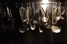 A collection of various kitchen utensils hanging from hooks on a metal rack. The utensils include a whisk, ladles, spatula, and grater, all made of metal. The arrangement is organized and illuminated by a soft, focused light creating a reflective sheen.