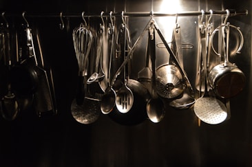A collection of various kitchen utensils hanging from hooks on a metal rack. The utensils include a whisk, ladles, spatula, and grater, all made of metal. The arrangement is organized and illuminated by a soft, focused light creating a reflective sheen.