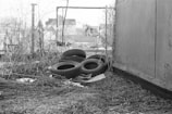 A black and white photograph of an outdoor area featuring discarded tires stacked on a sheet. Surrounding the tires are dry grass and twigs, with a wooden fence seen in the background. The setting appears neglected and cluttered, hinting at an urban or industrial environment.