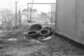 A black and white photograph of an outdoor area featuring discarded tires stacked on a sheet. Surrounding the tires are dry grass and twigs, with a wooden fence seen in the background. The setting appears neglected and cluttered, hinting at an urban or industrial environment.