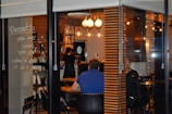 A cozy cafe interior is visible through a glass window. Inside, a few people are seated at wooden tables, and a barista is working behind a counter. Warm lighting from hanging light bulbs creates an inviting atmosphere. Menu items are handwritten on the glass, adding a unique touch to the decor.