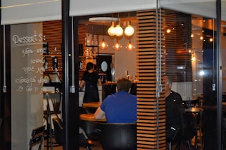 A cozy cafe interior is visible through a glass window. Inside, a few people are seated at wooden tables, and a barista is working behind a counter. Warm lighting from hanging light bulbs creates an inviting atmosphere. Menu items are handwritten on the glass, adding a unique touch to the decor.
