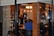 A cozy cafe interior is visible through a glass window. Inside, a few people are seated at wooden tables, and a barista is working behind a counter. Warm lighting from hanging light bulbs creates an inviting atmosphere. Menu items are handwritten on the glass, adding a unique touch to the decor.