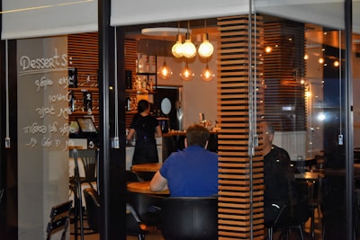 A cozy cafe interior is visible through a glass window. Inside, a few people are seated at wooden tables, and a barista is working behind a counter. Warm lighting from hanging light bulbs creates an inviting atmosphere. Menu items are handwritten on the glass, adding a unique touch to the decor.