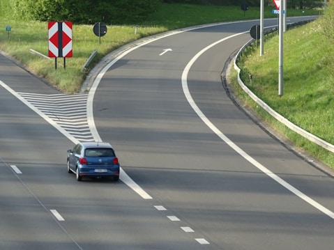 A blue car is driving on a highway with clearly marked white lines. The road curves to the right and there is green grass along the roadside. Various road signs are present, including a red and white chevron sign and directional signs.