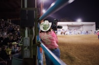 A photographer wearing a cowboy hat and pink shirt with intricate embroidery stands by the fence, holding a camera with a large lens, capturing events in an indoor arena. The scene is lively, with spectators sitting in the bleachers and people scattered near the center of the dirt arena.