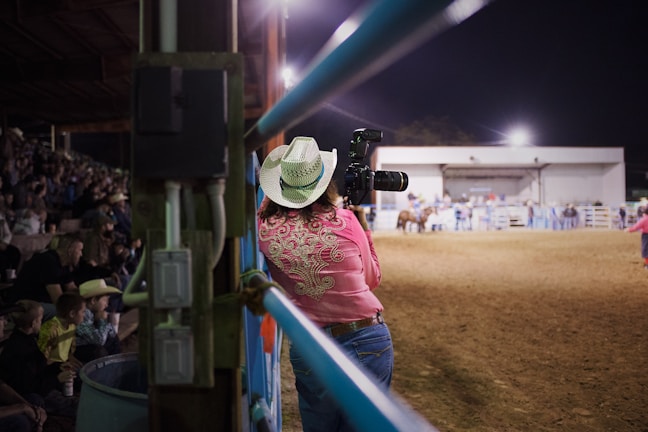 A photographer wearing a cowboy hat and pink shirt with intricate embroidery stands by the fence, holding a camera with a large lens, capturing events in an indoor arena. The scene is lively, with spectators sitting in the bleachers and people scattered near the center of the dirt arena.