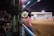 A photographer wearing a cowboy hat and pink shirt with intricate embroidery stands by the fence, holding a camera with a large lens, capturing events in an indoor arena. The scene is lively, with spectators sitting in the bleachers and people scattered near the center of the dirt arena.