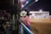 A photographer wearing a cowboy hat and pink shirt with intricate embroidery stands by the fence, holding a camera with a large lens, capturing events in an indoor arena. The scene is lively, with spectators sitting in the bleachers and people scattered near the center of the dirt arena.