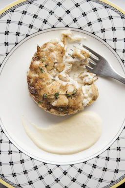 A close-up of a golden crab cake on a white plate garnished with lemon wedges and fresh herbs.