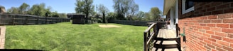 A spacious backyard with a well-maintained lawn surrounded by a wooden fence. A small wooden shed is situated near the center. The right side features a brick house with a small wooden deck and steps leading to the yard. Trees and a clear blue sky are visible in the background.