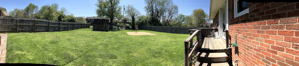 A spacious backyard with a well-maintained lawn surrounded by a wooden fence. A small wooden shed is situated near the center. The right side features a brick house with a small wooden deck and steps leading to the yard. Trees and a clear blue sky are visible in the background.