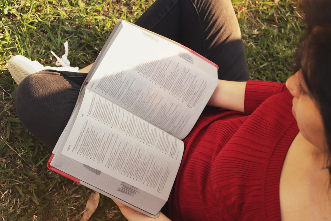 person in red long sleeve shirt and black pants sitting on green grass field reading book