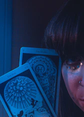 A close-up of a person's face partially visible, adorned with small star decorations near the eye and a nose ring. Two tarot cards, 'The Sun' and 'The Moon,' are prominently displayed in front of the face. The scene is enveloped in a mystical blue light.