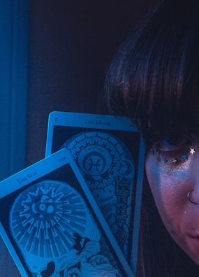 A close-up of a person's face partially visible, adorned with small star decorations near the eye and a nose ring. Two tarot cards, 'The Sun' and 'The Moon,' are prominently displayed in front of the face. The scene is enveloped in a mystical blue light.