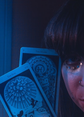 A close-up of a person's face partially visible, adorned with small star decorations near the eye and a nose ring. Two tarot cards, 'The Sun' and 'The Moon,' are prominently displayed in front of the face. The scene is enveloped in a mystical blue light.