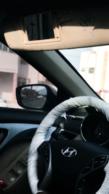 A view of a car interior is visible, focusing on the steering wheel with a wrapped cover displaying the Hyundai logo. The dashboard and interior door controls are also visible. Sunlight casts shadows, enhancing the vehicle's features. The outside street view can be seen through the side mirror and window, featuring nearby buildings.