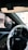 A view of a car interior is visible, focusing on the steering wheel with a wrapped cover displaying the Hyundai logo. The dashboard and interior door controls are also visible. Sunlight casts shadows, enhancing the vehicle's features. The outside street view can be seen through the side mirror and window, featuring nearby buildings.