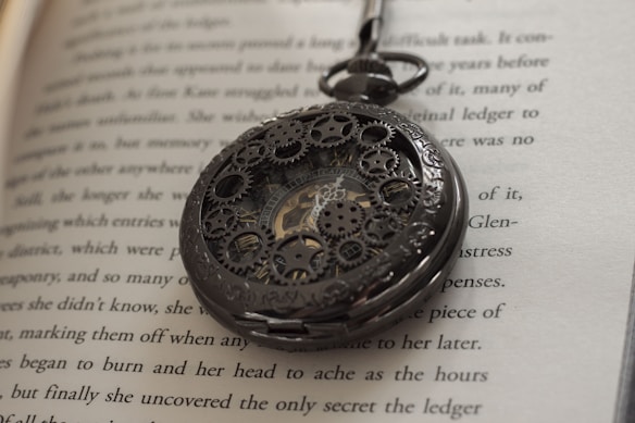 An ornate pocket watch with intricate gear and mechanical details lies atop an open book with text visible on the pages. The focus is on the watch, highlighting its design and craftsmanship.