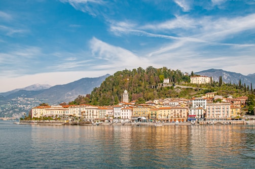A picturesque lakeside town with colorful buildings along the waterfront. The architecture is a mix of historical and traditional styles. Behind the town, a lush green hill rises, covered with trees and a few scattered villas. Majestic mountains stand in the background under a sky with wispy clouds.