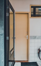 A narrow entryway featuring a closed beige door with silver hardware. The area to the left includes a glass window, reflecting part of the scene. On the right, a section of white tiles with a gray border is visible, preceded by a partial view of a bicycle wheel. Above the door, there is a small window with horizontal panes.